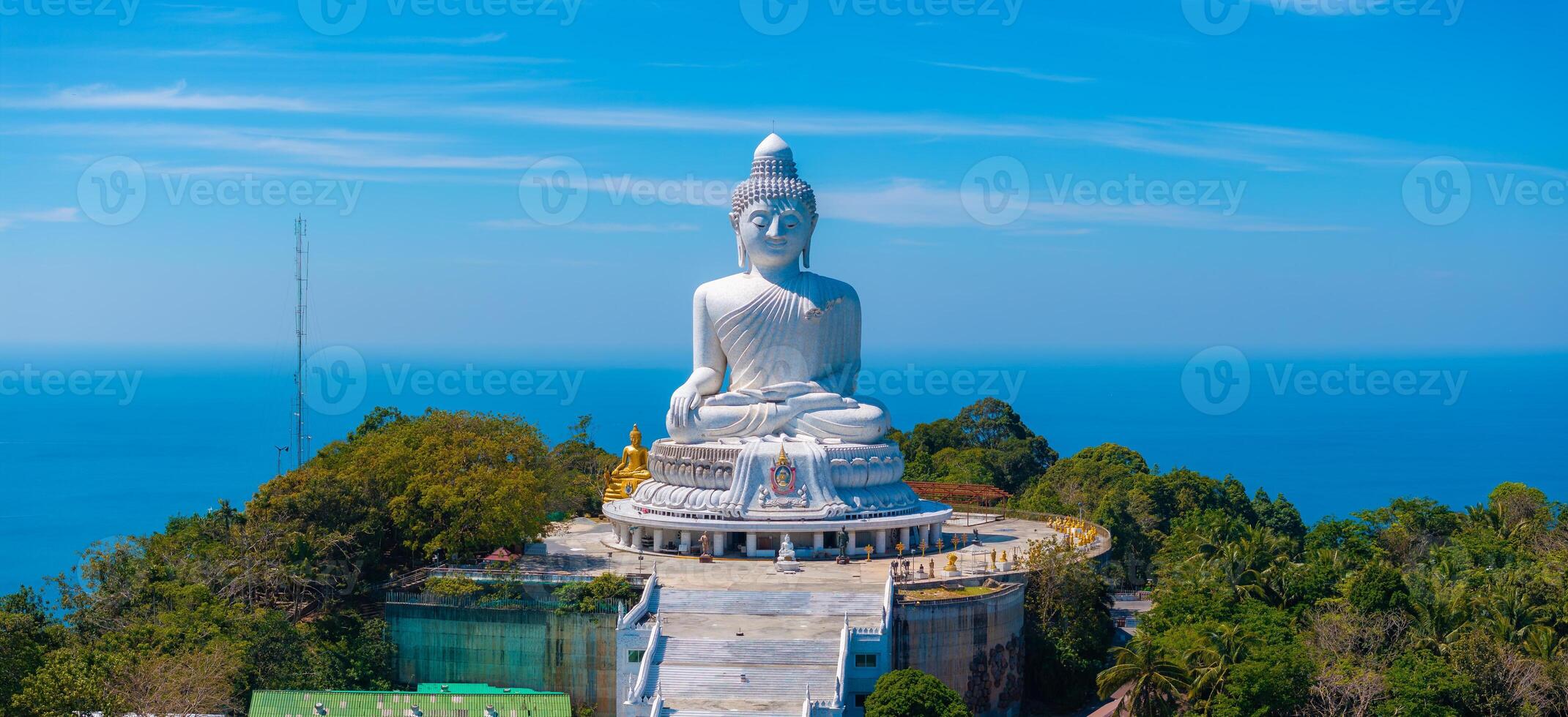 The iconic white marble Big Buddha statue sits in a meditative pose atop a hill in Phuket, Thailand, surrounded by greenery and ocean views under a clear blue sky. photo