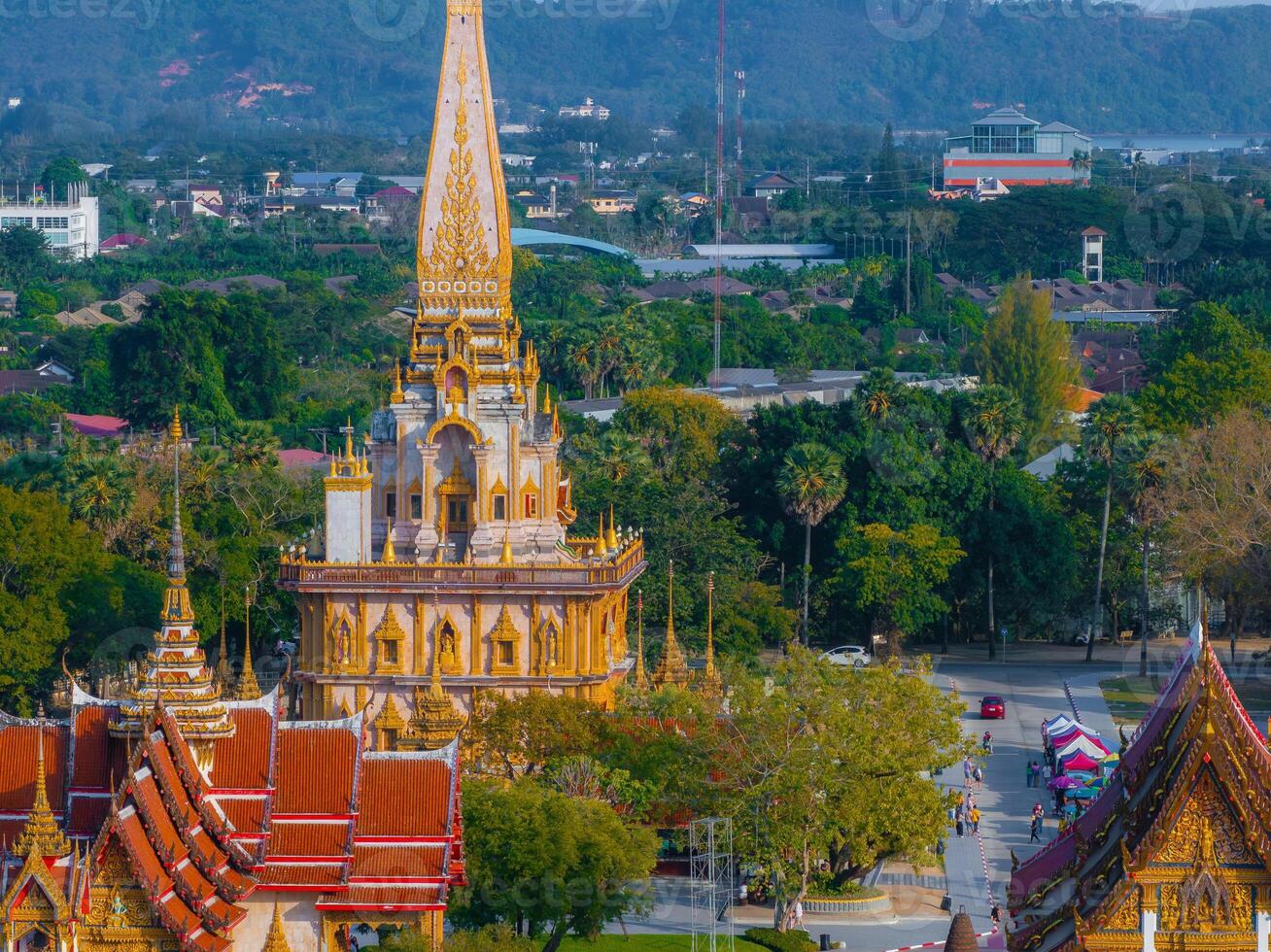The ornate Wat Chalong temple in Phuket, Thailand, features a golden stupa, red and gold roofs, lush greenery, and distant hills in the background. photo