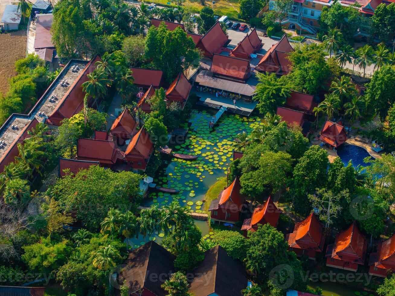 Aerial view of red roofed Thai buildings with pointed gables, surrounded by lush greenery and a large lotus pond filled with lily pads in Phuket, Thailand. photo
