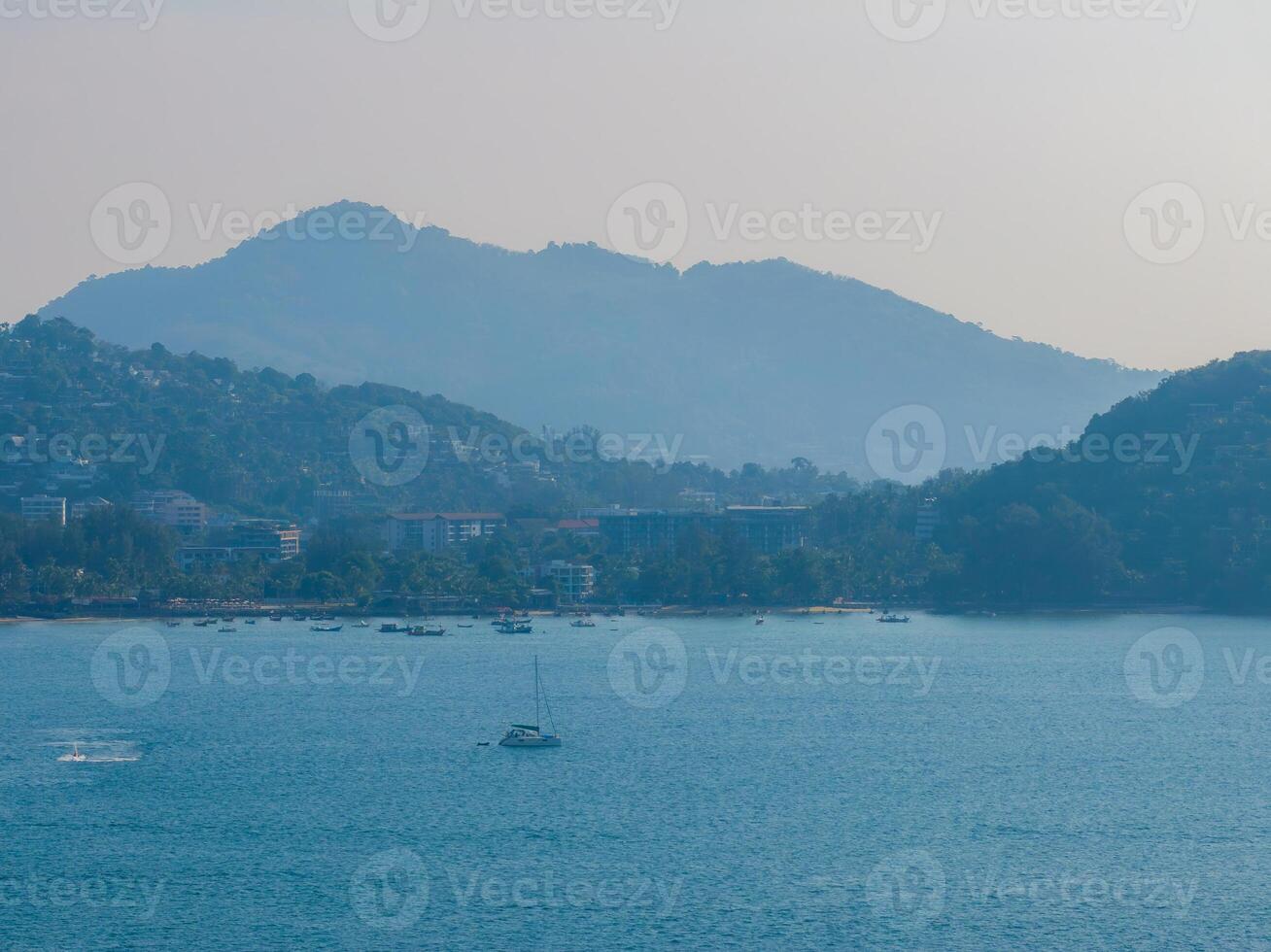 Calm blue waters with a single sailboat, lush green hills in the background, and a small coastal town nestled along the shoreline in Phuket. photo