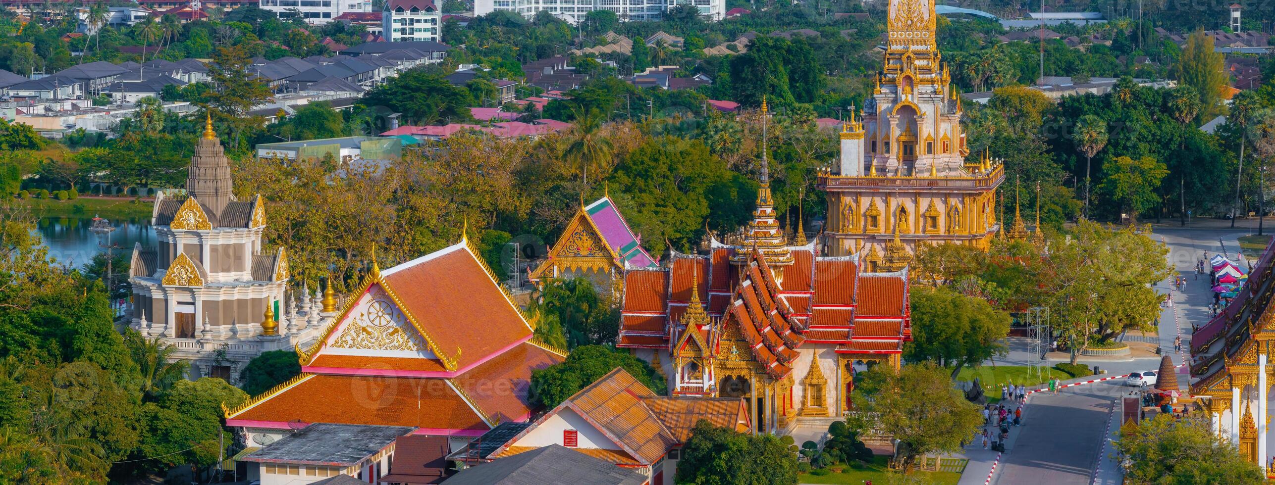 Wat Chalong's golden stupa stands amidst red and gold roofed structures, surrounded by greenery and urban buildings in Phuket, Thailand. photo