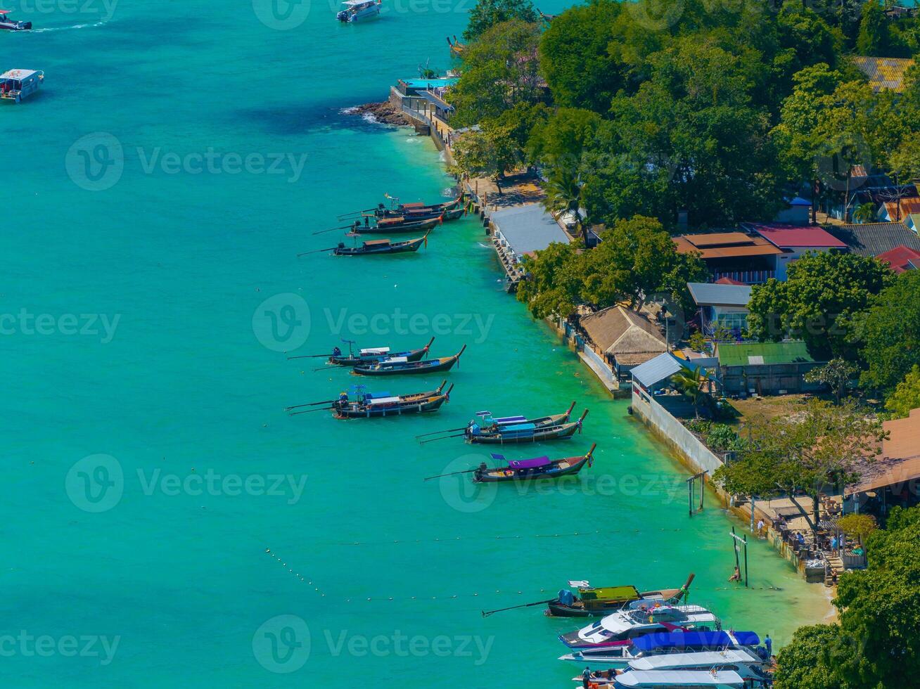 Turquoise waters and long tail boats line the shoreline of Phi Phi Islands, with small buildings and lush greenery in a vibrant coastal setting. photo