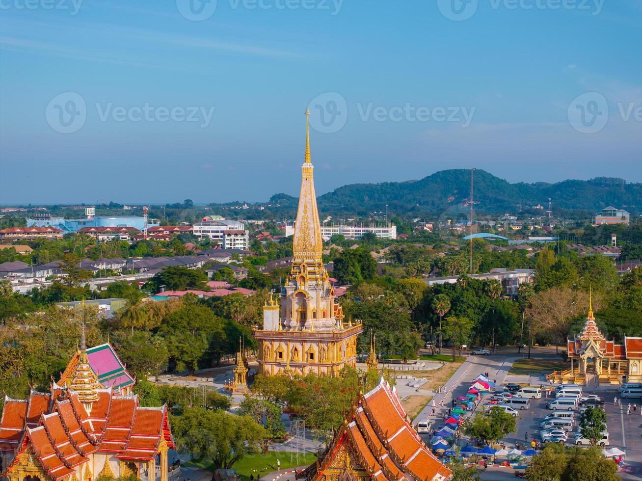 Wat Chalong temple in Phuket, Thailand, features a golden spire, intricate red and gold roofs, lush greenery, distant hills, and a clear blue sky. photo
