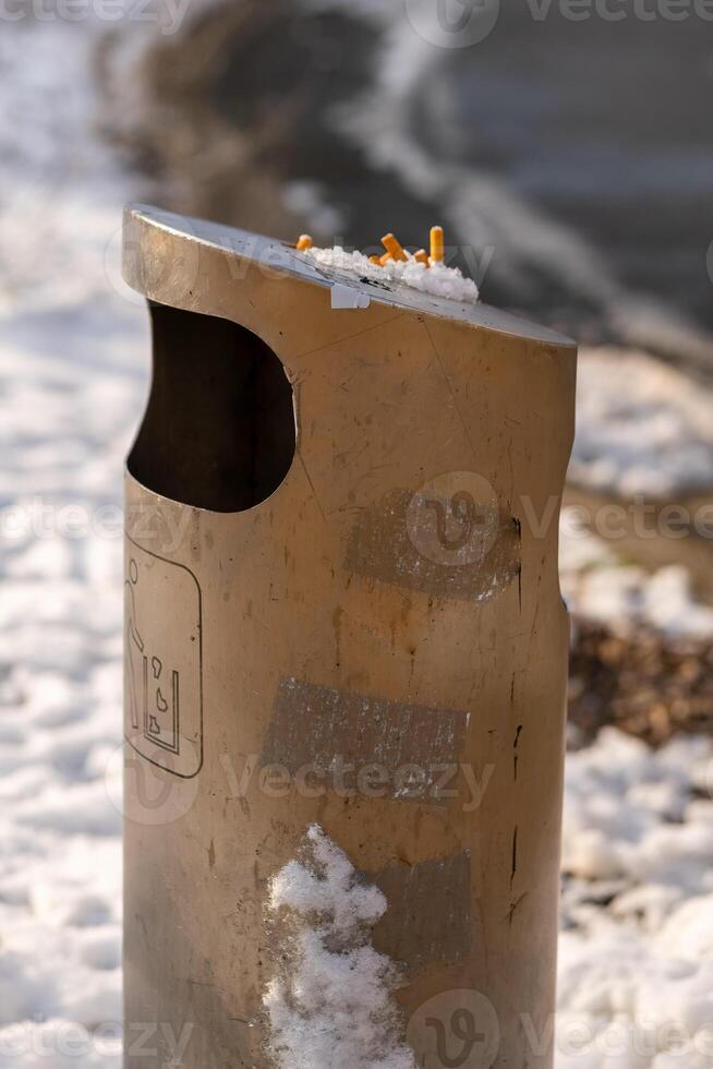 Discarded cigarette butts pile on top of a metal trash can near snow-covered ground in a cold location photo