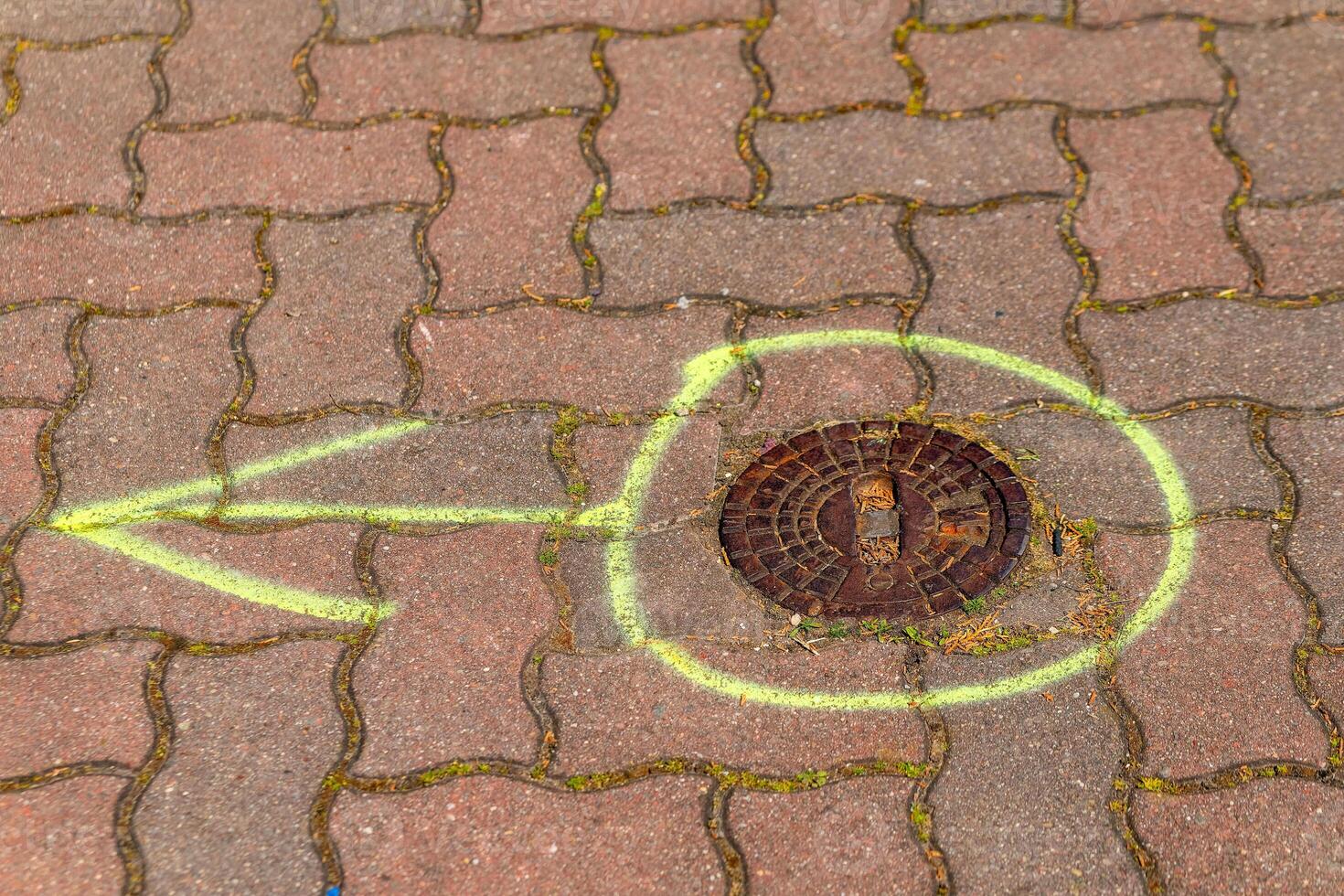 Bright yellow arrow and circle marked on pavement leading to a manhole cover surrounded by moss and paved stones photo