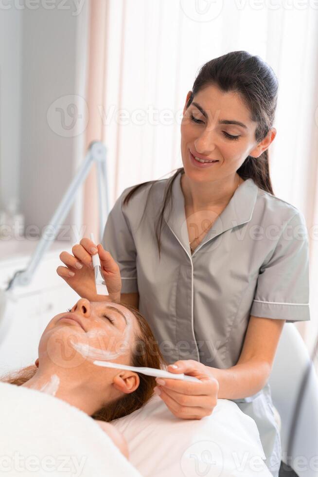 Happy therapist applying face mask to woman with brush at salon photo