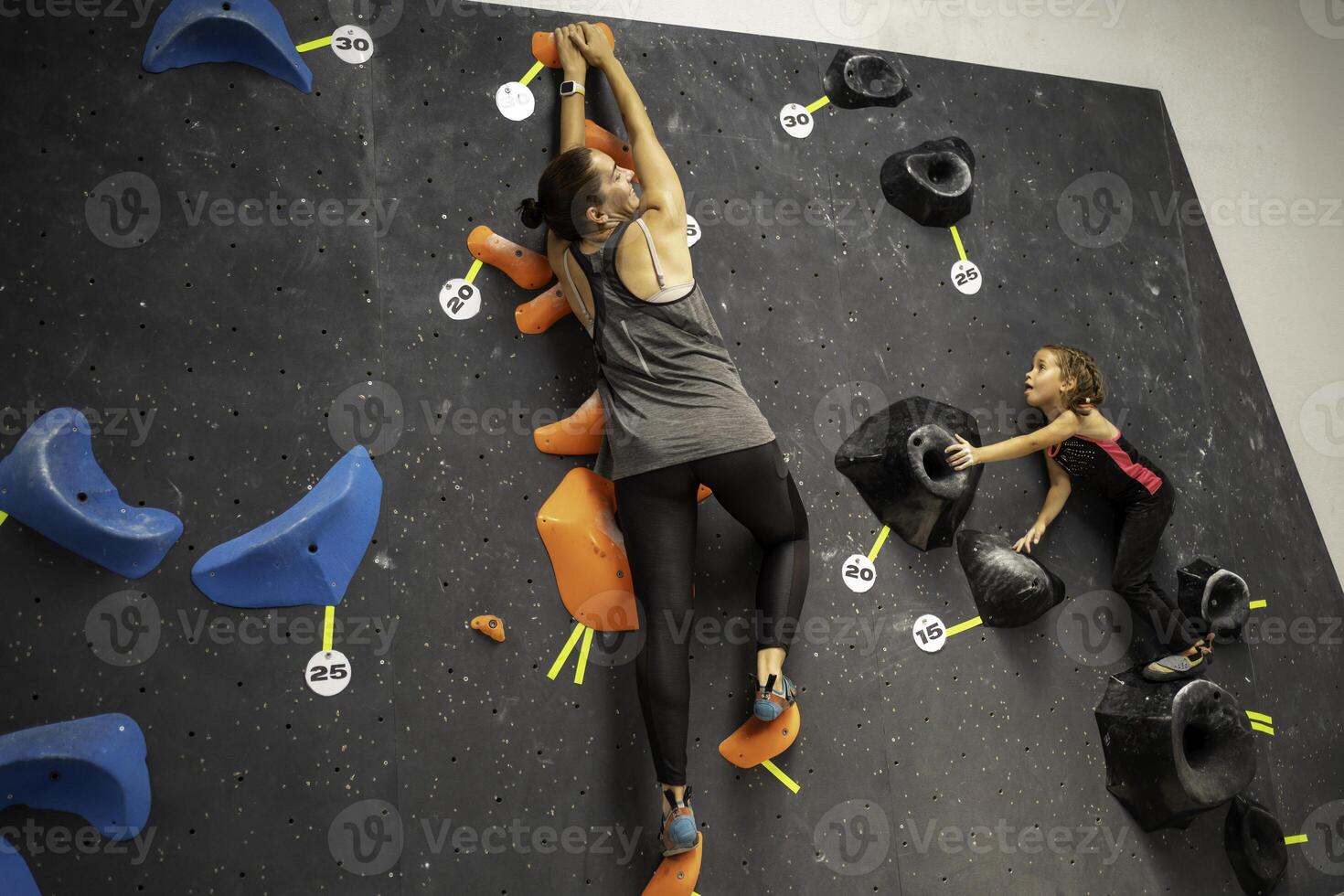 Courageous girl climbing artificial rock wall with mother at gym photo