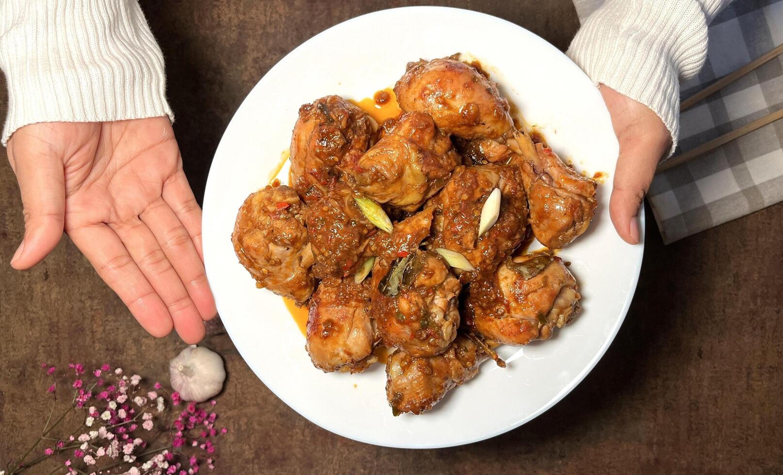 Woman hand is holding and showing a plate of delicious looking chicken drumsticks in a rich sauce, garnished with green onions and garlic photo