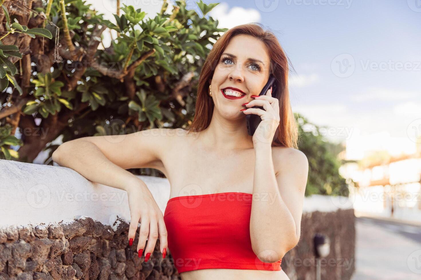 An attractive happy woman in a red top is having a phone call photo