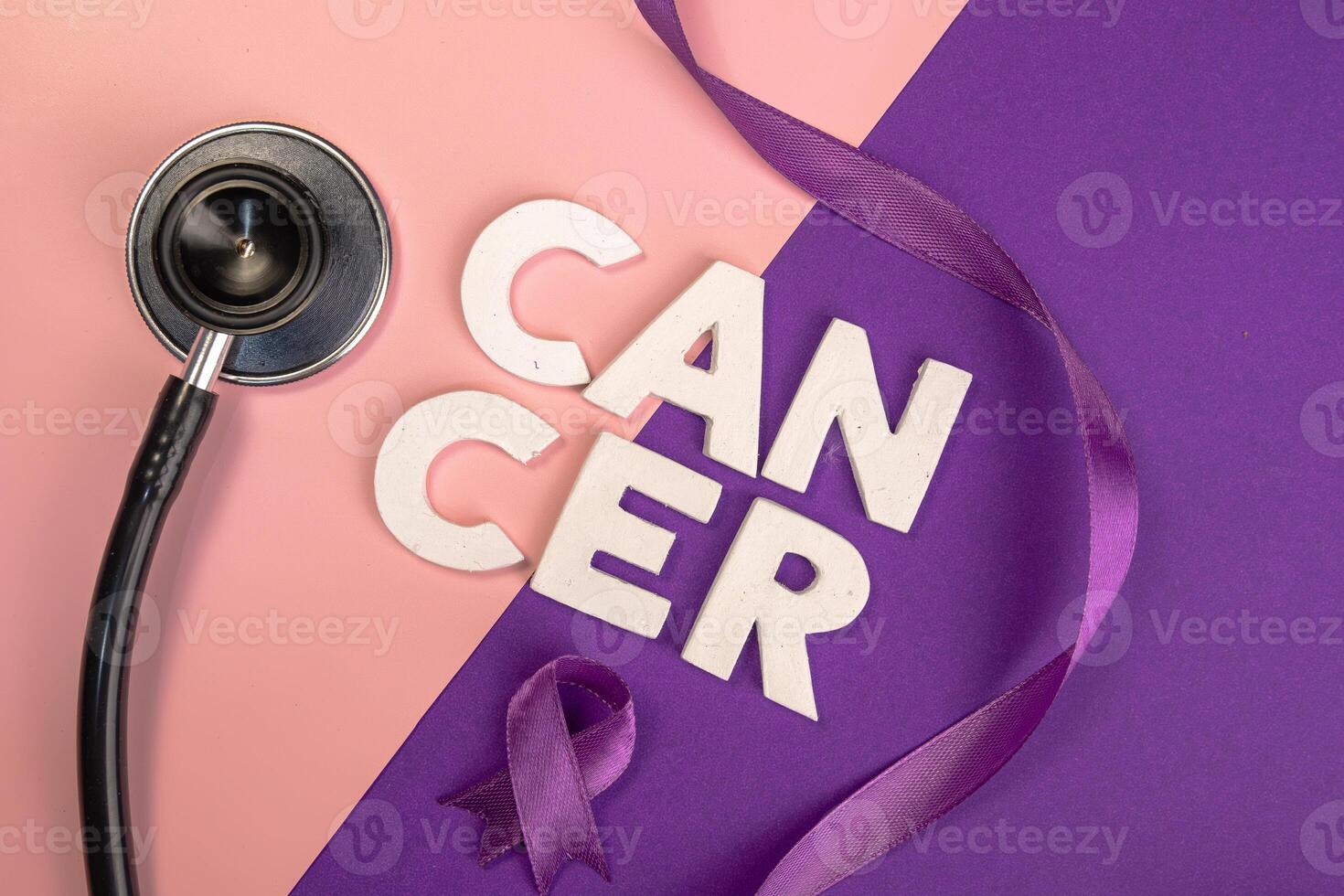 A stethoscope lies near the word CANCER spelled out in blocks next to a purple ribbon. World cancer day concept photo