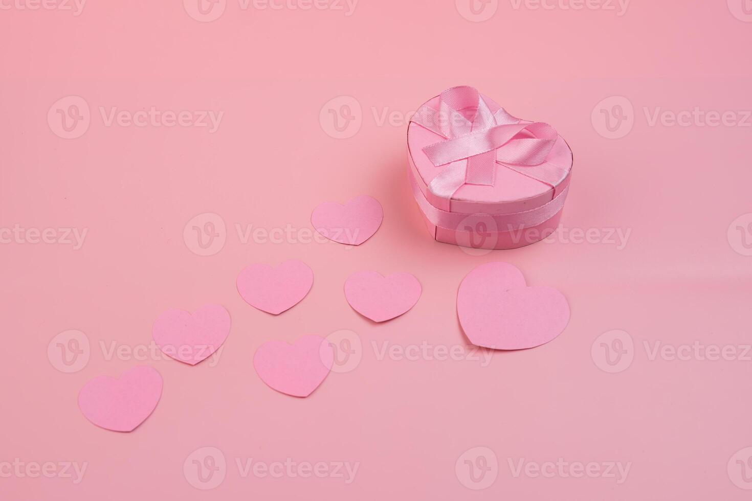 A heart shaped gift box with a pink ribbon sits beside several pink paper hearts. valentines day concept photo
