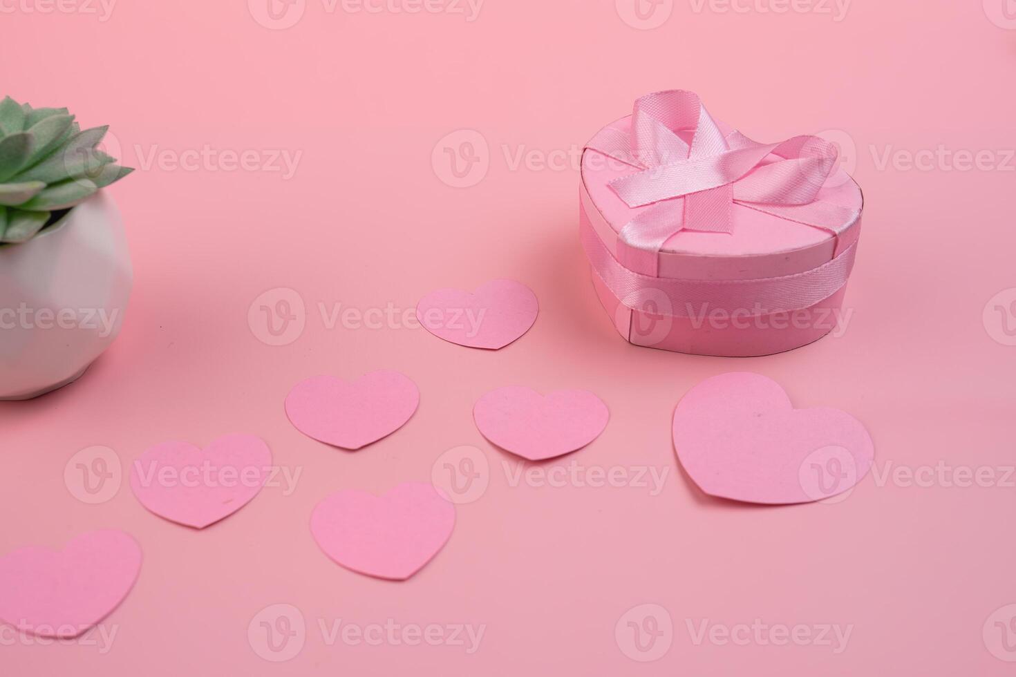 A heart shaped gift box with a pink ribbon sits next to a trail of pink paper hearts and a succulent. valentines day concept photo