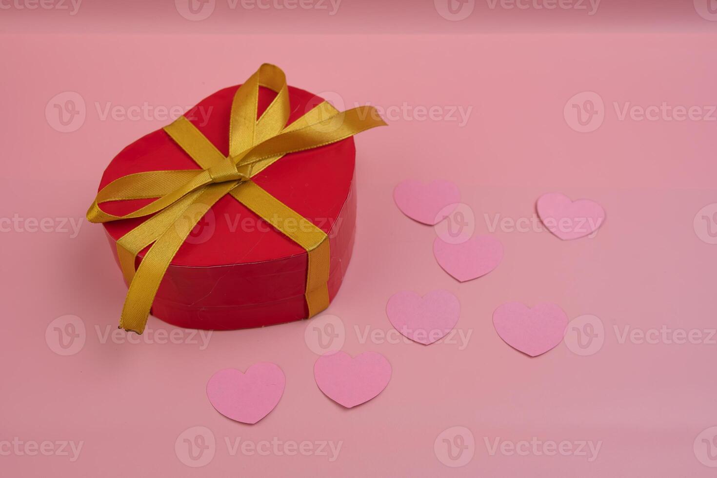 A heart shaped red gift box with a gold ribbon sits on a pink surface with small pink hearts scattered around it photo