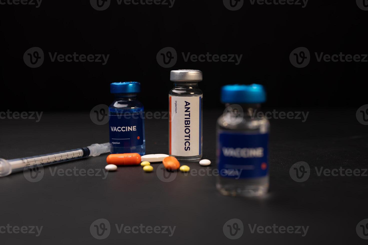 Two vaccine vials an antibiotics vial a syringe and pills are sitting on a dark surface. Health and medical concept background photo