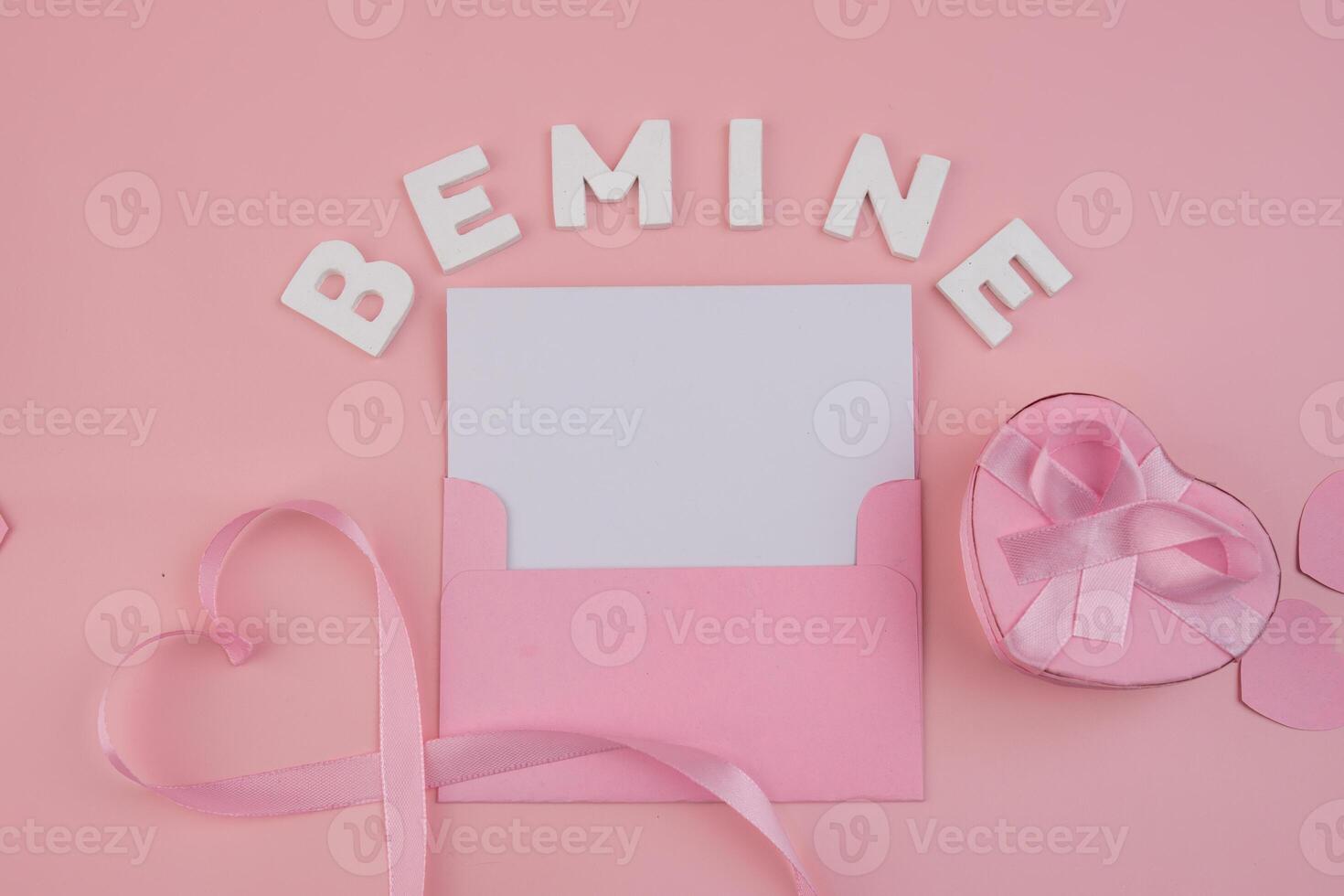 A pink envelope containing a blank card surrounded by the words Be Mine a pink ribbon shaped as a heart and a pink heart shaped gift box photo