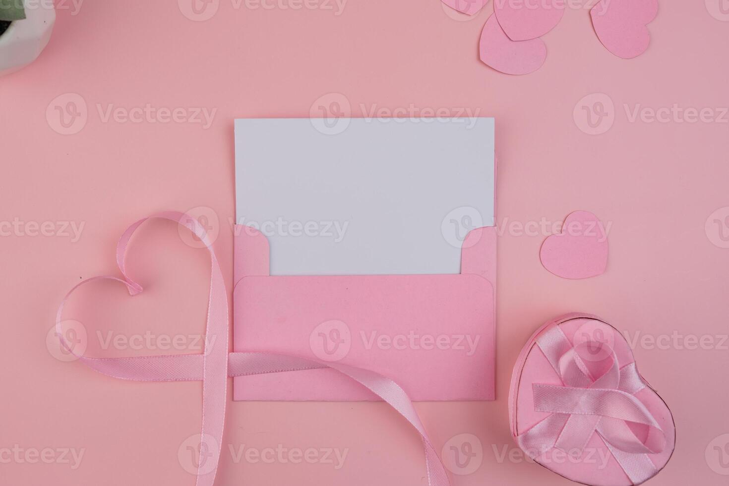 A pink envelope containing a blank card with a ribbon forming a heart shape next to a heart shaped gift box. invitation mockup photo