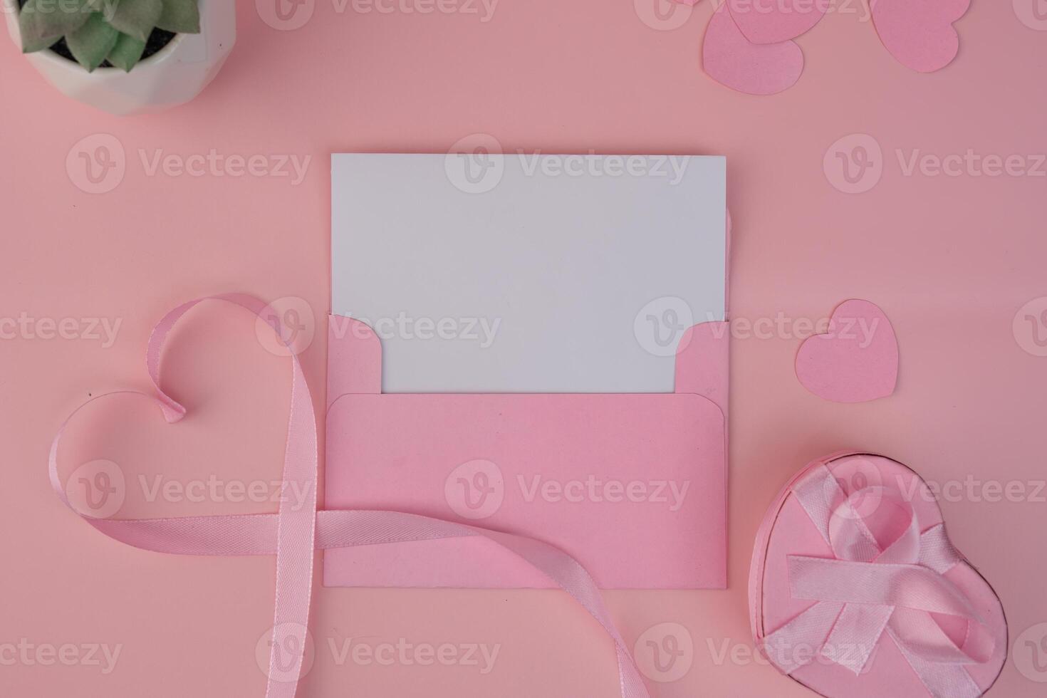 A pink envelope containing a blank card with a pink ribbon forming a heart shape next to a heart shaped gift box. invitation mockup design photo