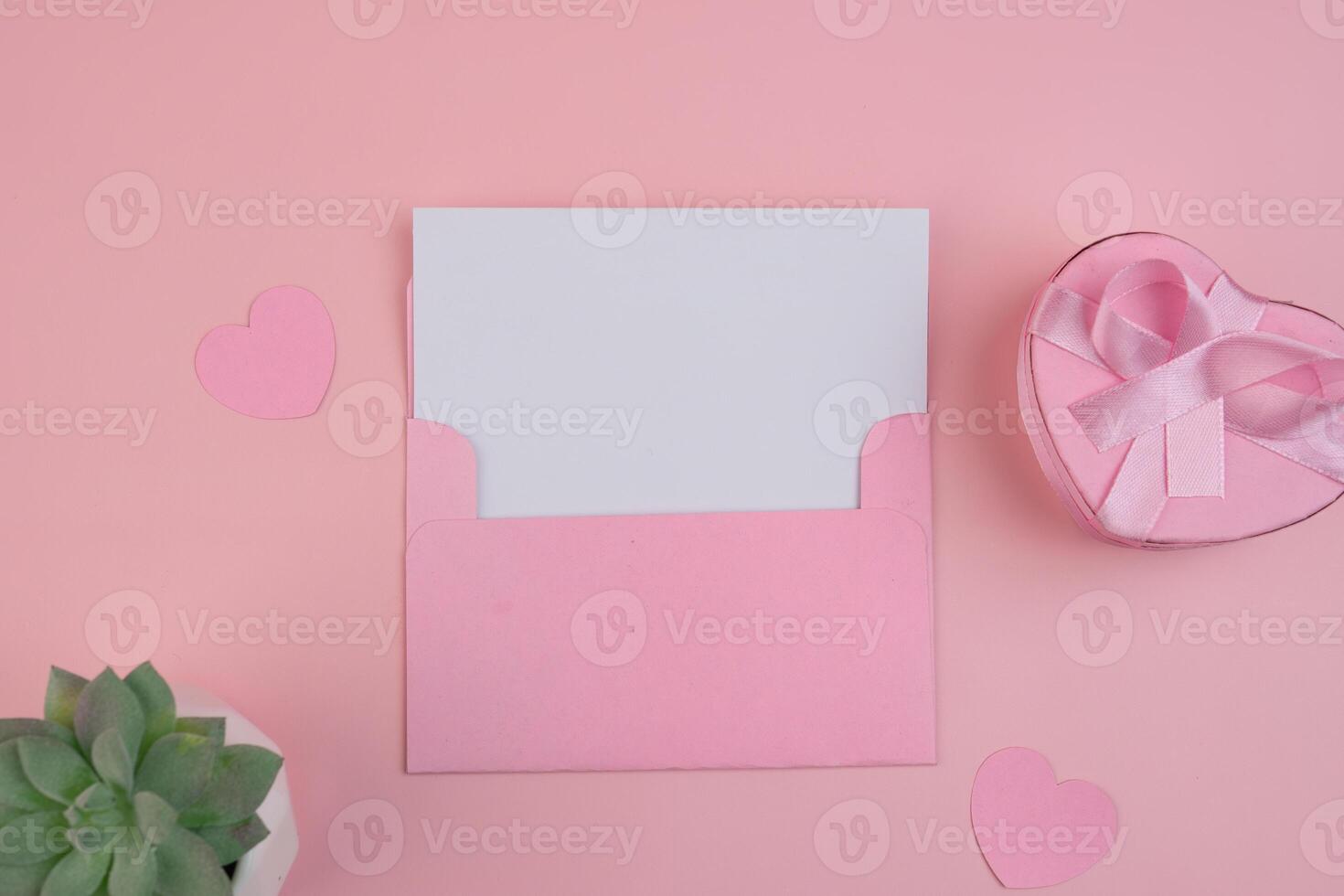 A pink envelope holding a blank white card next to a heart shaped gift box and paper hearts photo