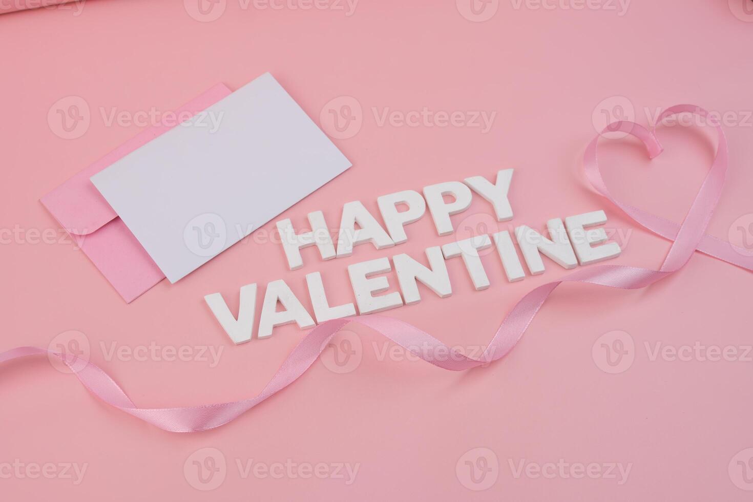 A pink ribbon forms a heart shape around the words Happy Valentine next to a blank card in a pink envelope photo