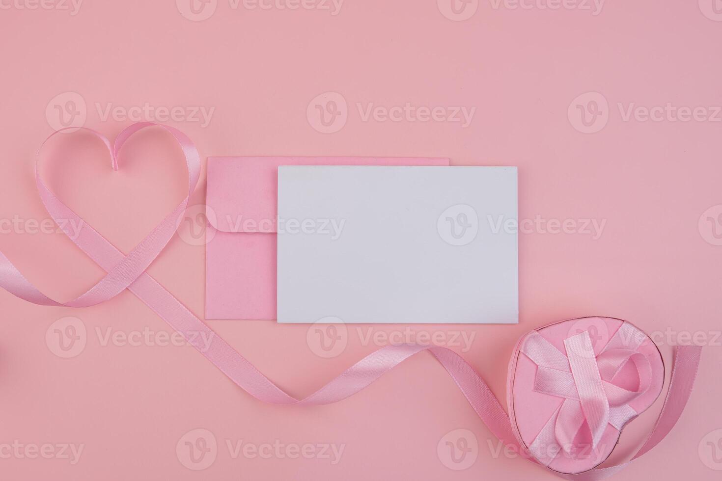 A pink ribbon forming a heart shape next to a pink envelope and a blank card with a heart shaped gift box tied with a ribbon photo