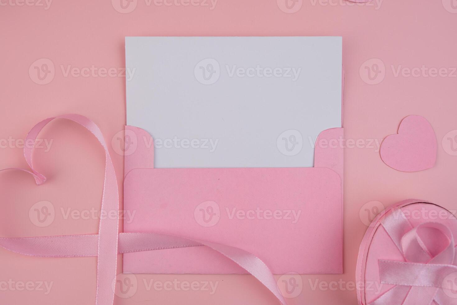 A pink envelope containing a blank white card next to a pink ribbon shaped as a heart and a pink gift box with a pink ribbon. invitation mockup photo