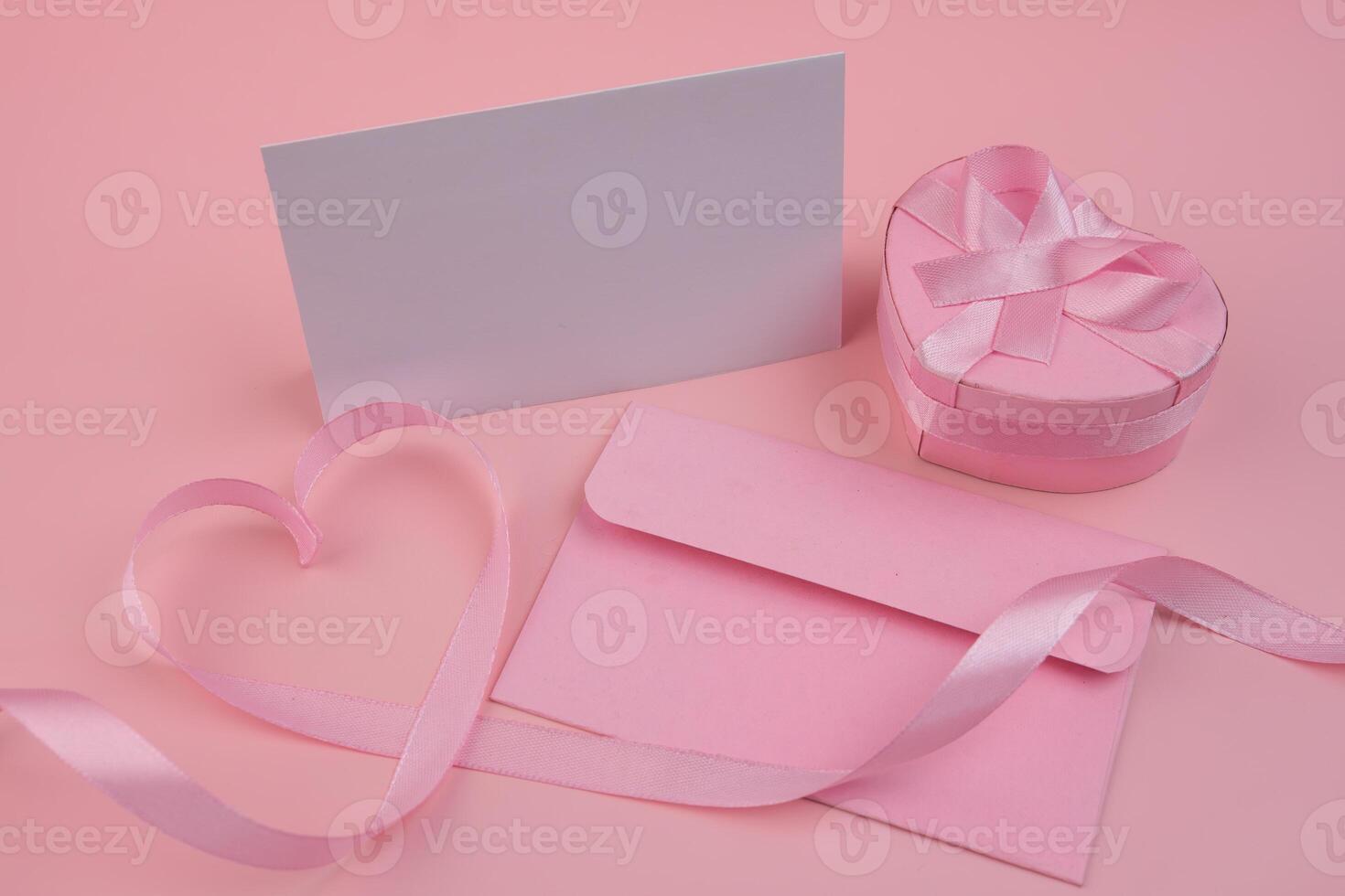 A pink ribbon forming a heart shape next to a pink envelope a blank card and a heart shaped gift box with a pink ribbon photo