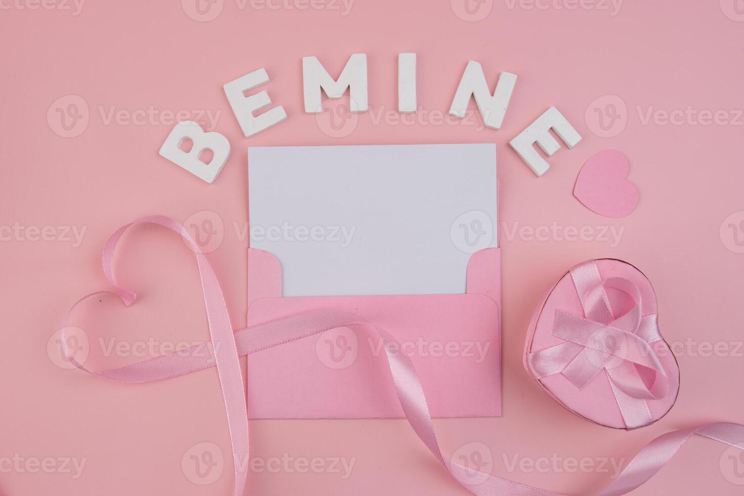 A pink envelope containing a blank card surrounded by a pink ribbon forming a heart a heart shaped gift box with a pink ribbon and the words Be Mine spelled out in white letters photo