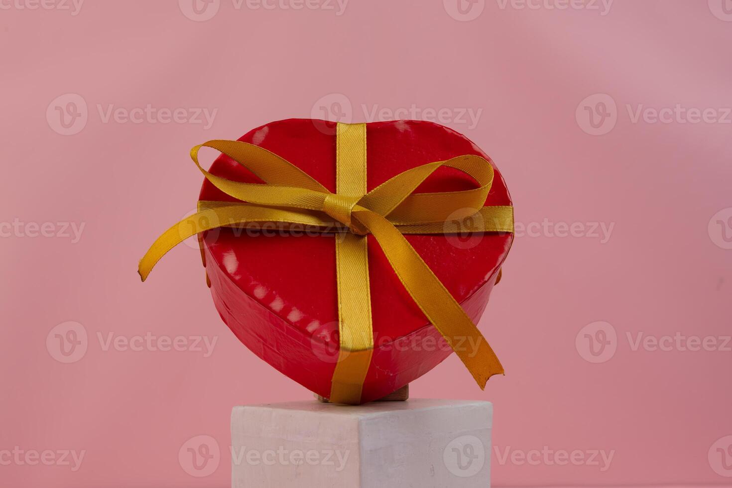 A heart shaped red gift box with a gold ribbon sits on a white cube photo