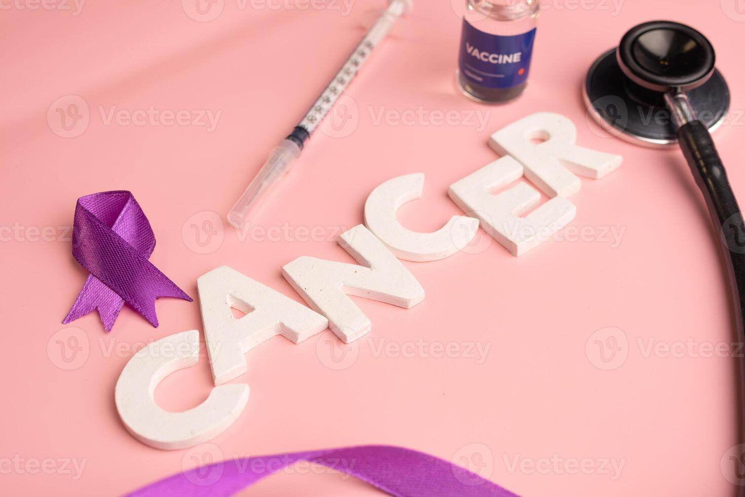 The word CANCER is spelled out with white letters alongside a purple ribbon a syringe a vaccine vial and a stethoscope. photo