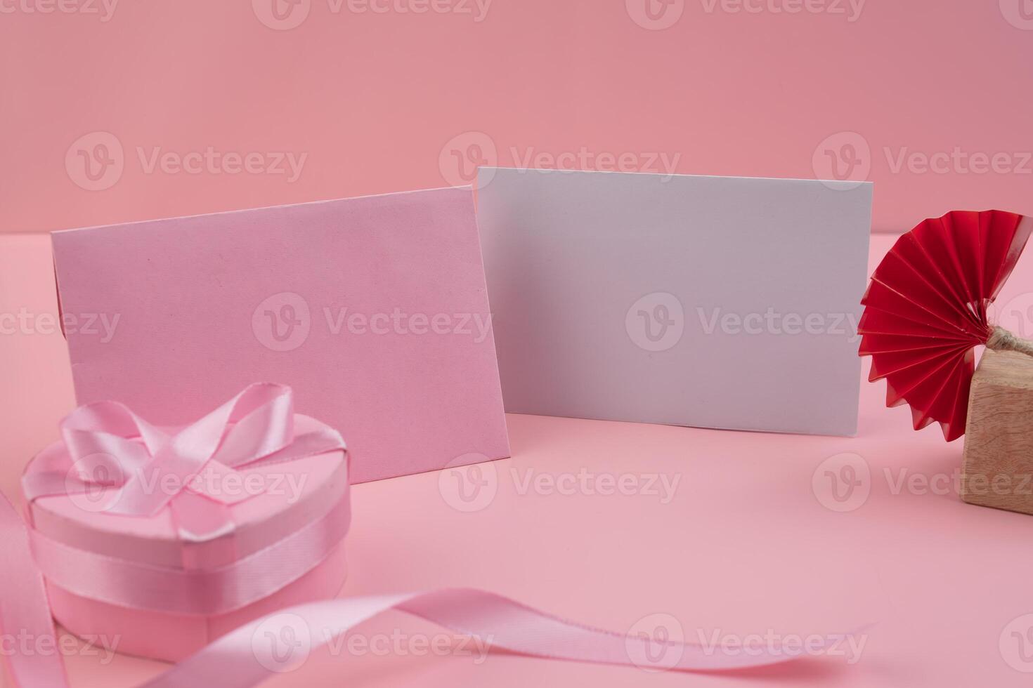 A pink heart shaped box with a pink ribbon sits next to a pink envelope and a blank card A red paper fan sits on a small wooden block photo