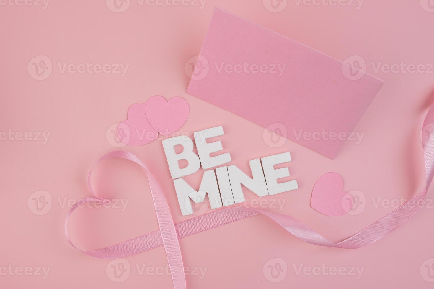 Pink hearts a ribbon forming a heart and letter blocks spelling Be Mine lay on a pink background next to a blank pink card photo