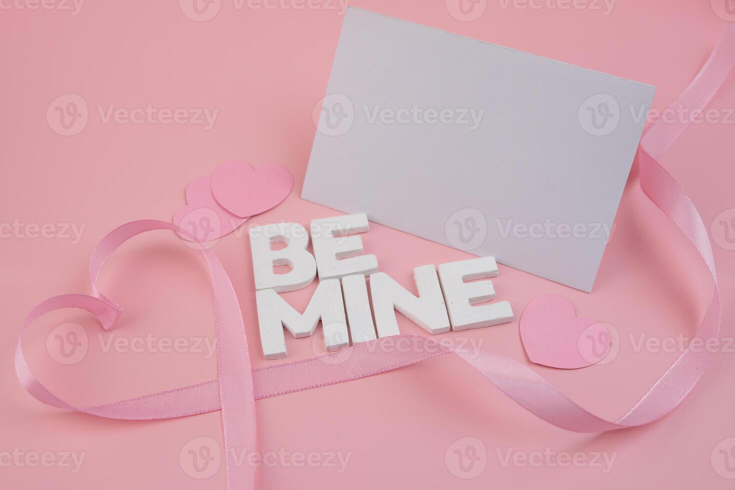 Pink ribbon forming hearts around white block letters spelling Be Mine and pink paper hearts A blank card lies nearby photo