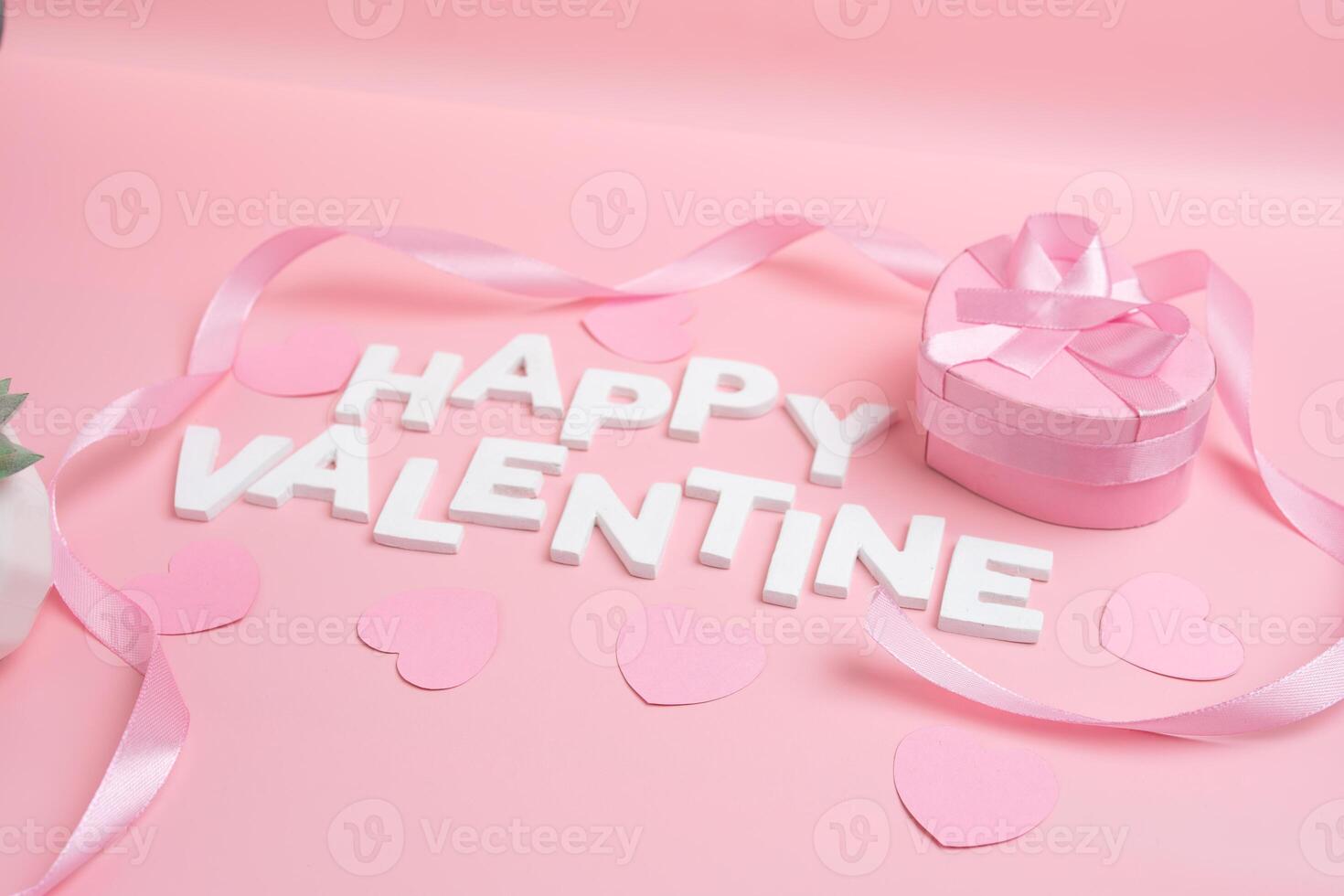 Happy Valentine message spelled out with white letters surrounded by pink hearts and ribbon next to a heart shaped gift box with a pink ribbon photo