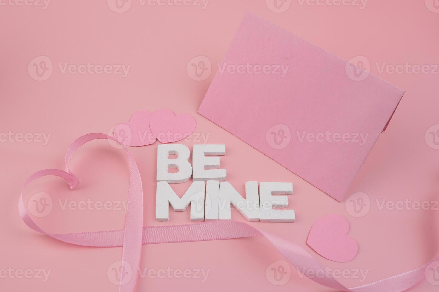 Pink ribbon forming a heart shape with small hearts wooden letters spelling Be Mine and a pink envelope photo
