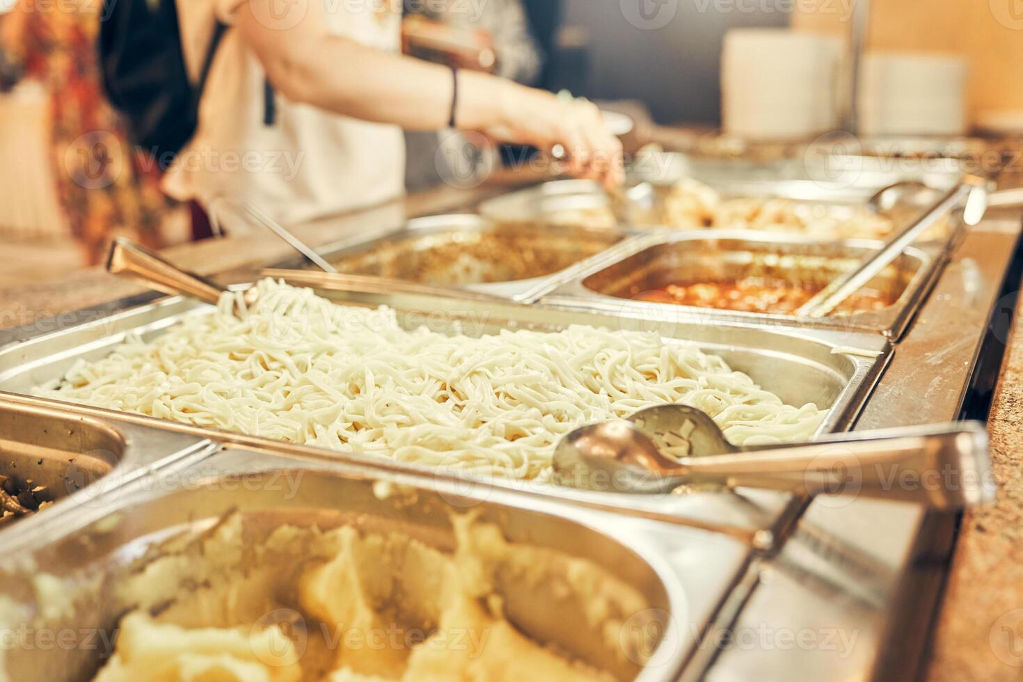 People are putting food on their plates. Self-service dining room. Close-up of whipped mashed potatoes in a metal tray next to spaghetti. photo