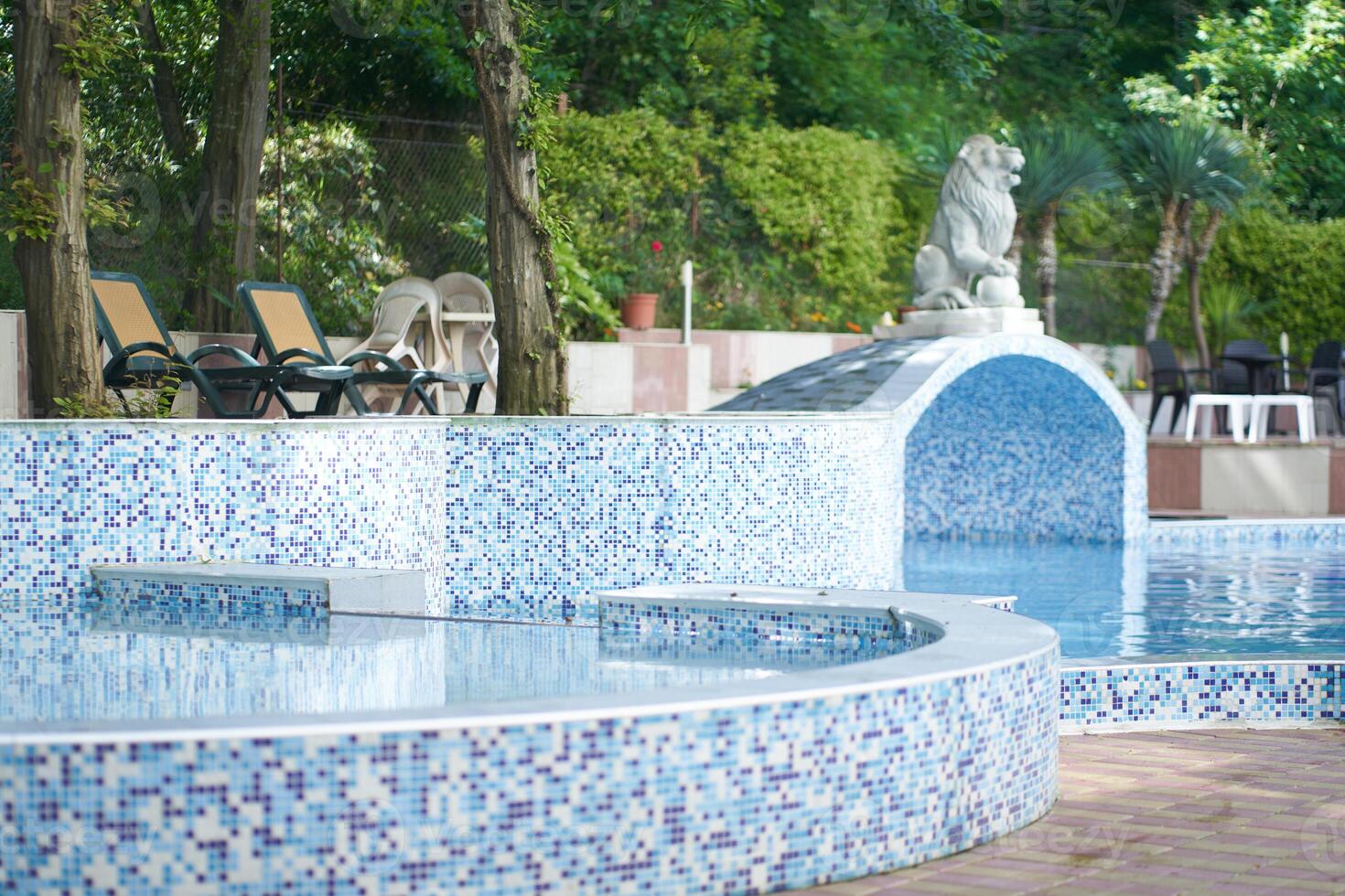 View of the hotel with swimming pool with mosaic tiles, sun loungers, a statue, a lion figure against the background of a forest, a park. Background with copy space. Concept of vacation, sunbathing photo