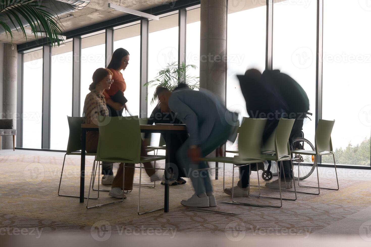 A diverse business team, working urgently around a conference table, collaborates on a meeting while their director, seated in a wheelchair, waits for their input photo