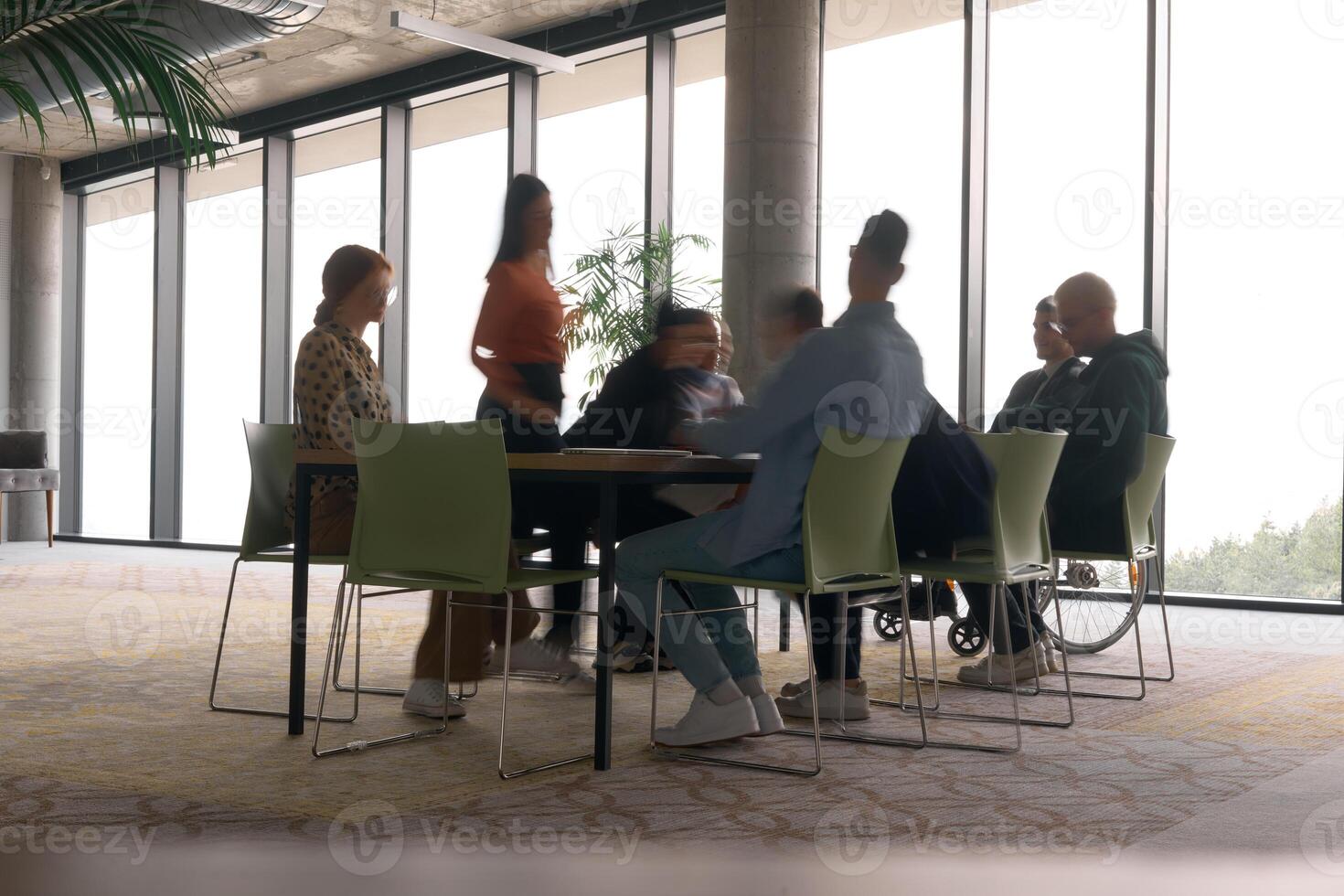 A diverse business team, working urgently around a conference table, collaborates on a meeting while their director, seated in a wheelchair, waits for their input photo