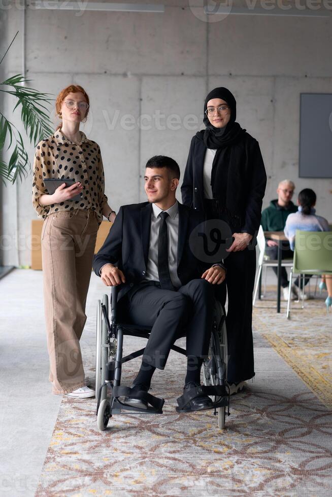 A diverse business team poses confidently, including a smiling director seated in a wheelchair, flanked by two professional female colleagues, one wearing a hijab and the other holding a tablet, in a photo