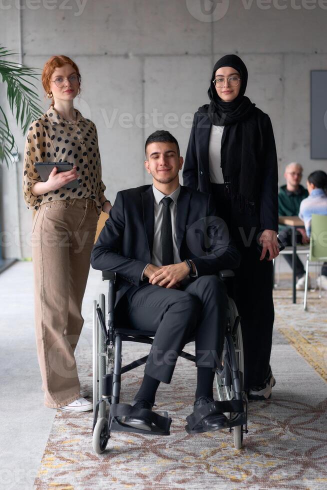 A diverse business team poses confidently, including a smiling director seated in a wheelchair, flanked by two professional female colleagues, one wearing a hijab and the other holding a tablet, in a photo