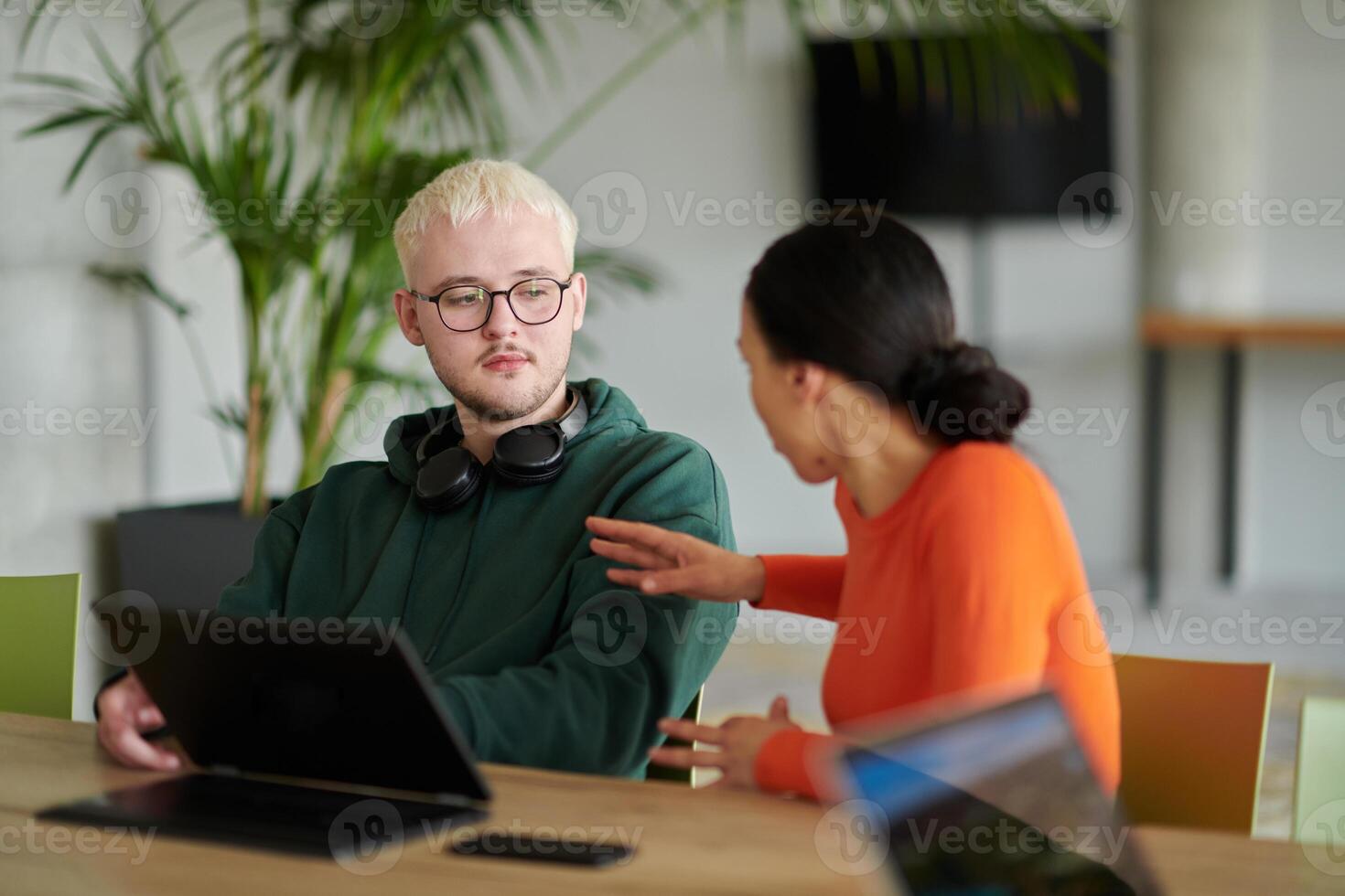 A blond businessman and his colleague sit in an office discussing work while using a laptop to collaborate on tasks photo