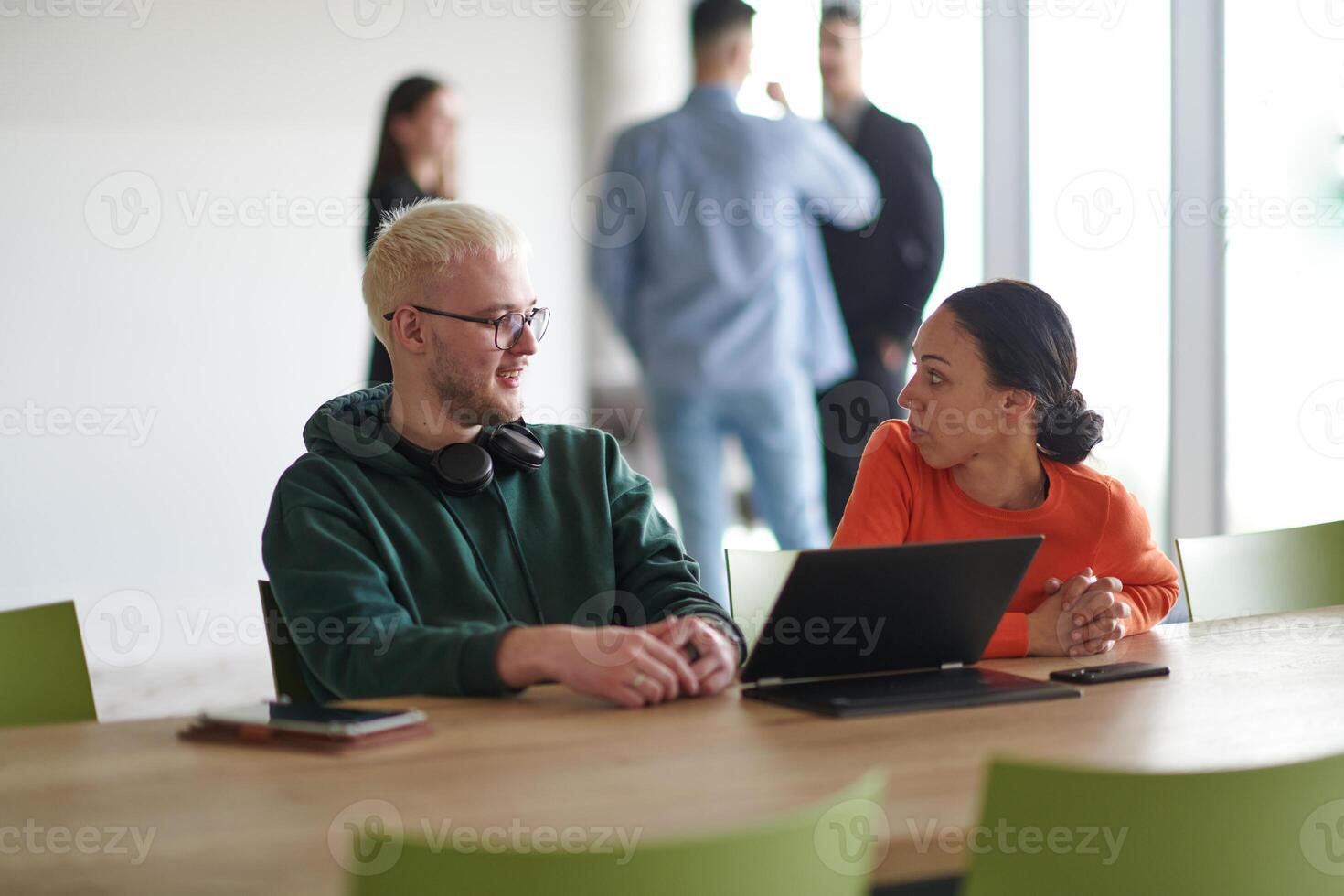 A blond businessman and his colleague sit in an office discussing work while using a laptop to collaborate on tasks photo