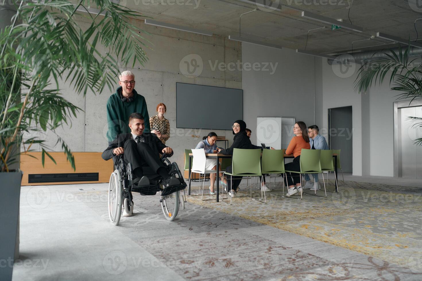 A diverse and multicultural business team collaborates in the office while their colleagues, including a director in a wheelchair and his friend, share a lighthearted moment, fostering an atmosphere photo