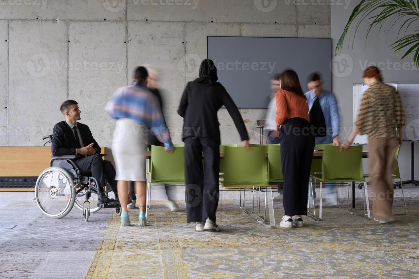 A diverse business team, working urgently around a conference table, collaborates on a meeting while their director, seated in a wheelchair, waits for their input photo