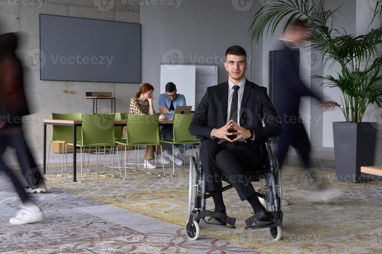 A businessman in a wheelchair navigating through a busy office, surrounded by his colleagues who are actively engaged in their work and collaboration photo