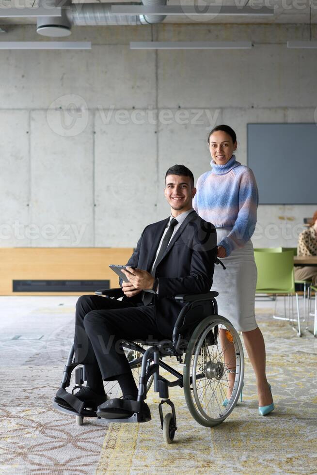 In a professional office setting, a businesswoman helping her director, who is sitting in a wheelchair and using a tablet, while their colleagues collaborate in the background. photo