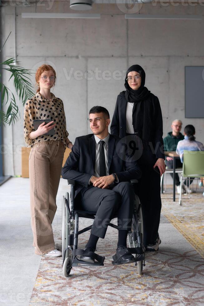 A diverse business team poses confidently, including a smiling director seated in a wheelchair, flanked by two professional female colleagues, one wearing a hijab and the other holding a tablet, in a photo