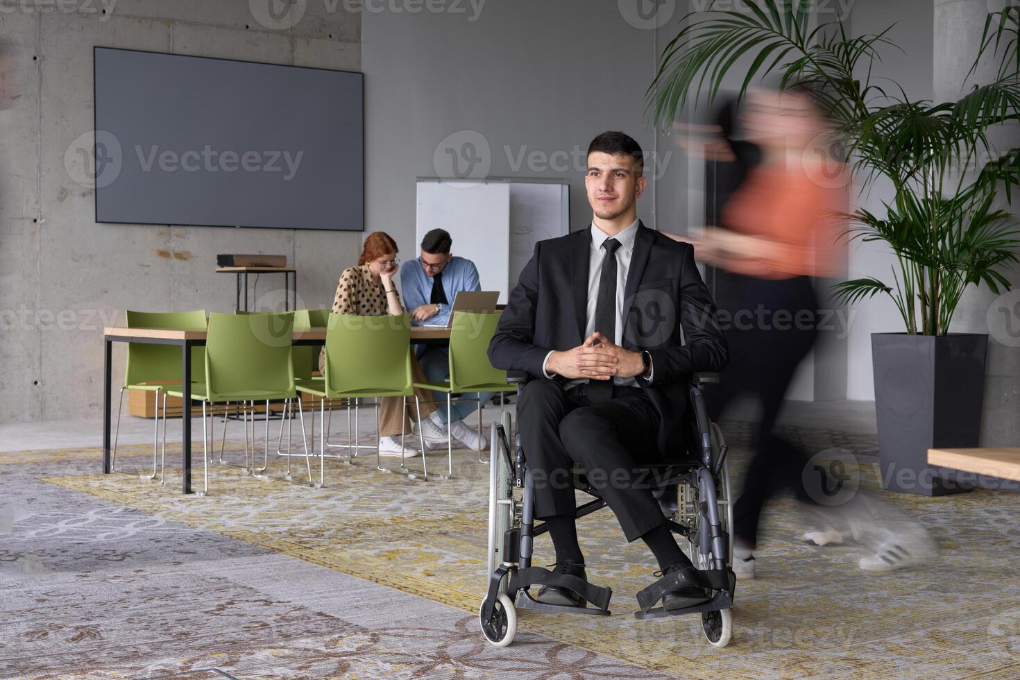 A businessman in a wheelchair navigating through a busy office, surrounded by his colleagues who are actively engaged in their work and collaboration photo