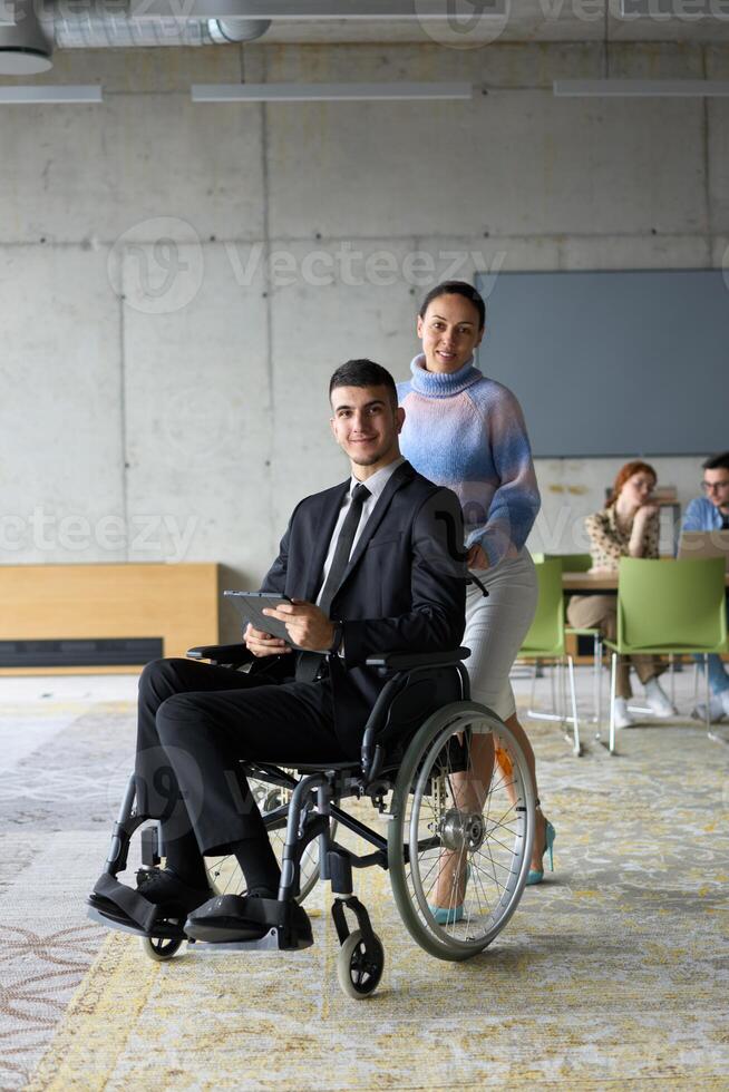In a professional office setting, a businesswoman helping her director, who is sitting in a wheelchair and using a tablet, while their colleagues collaborate in the background. photo