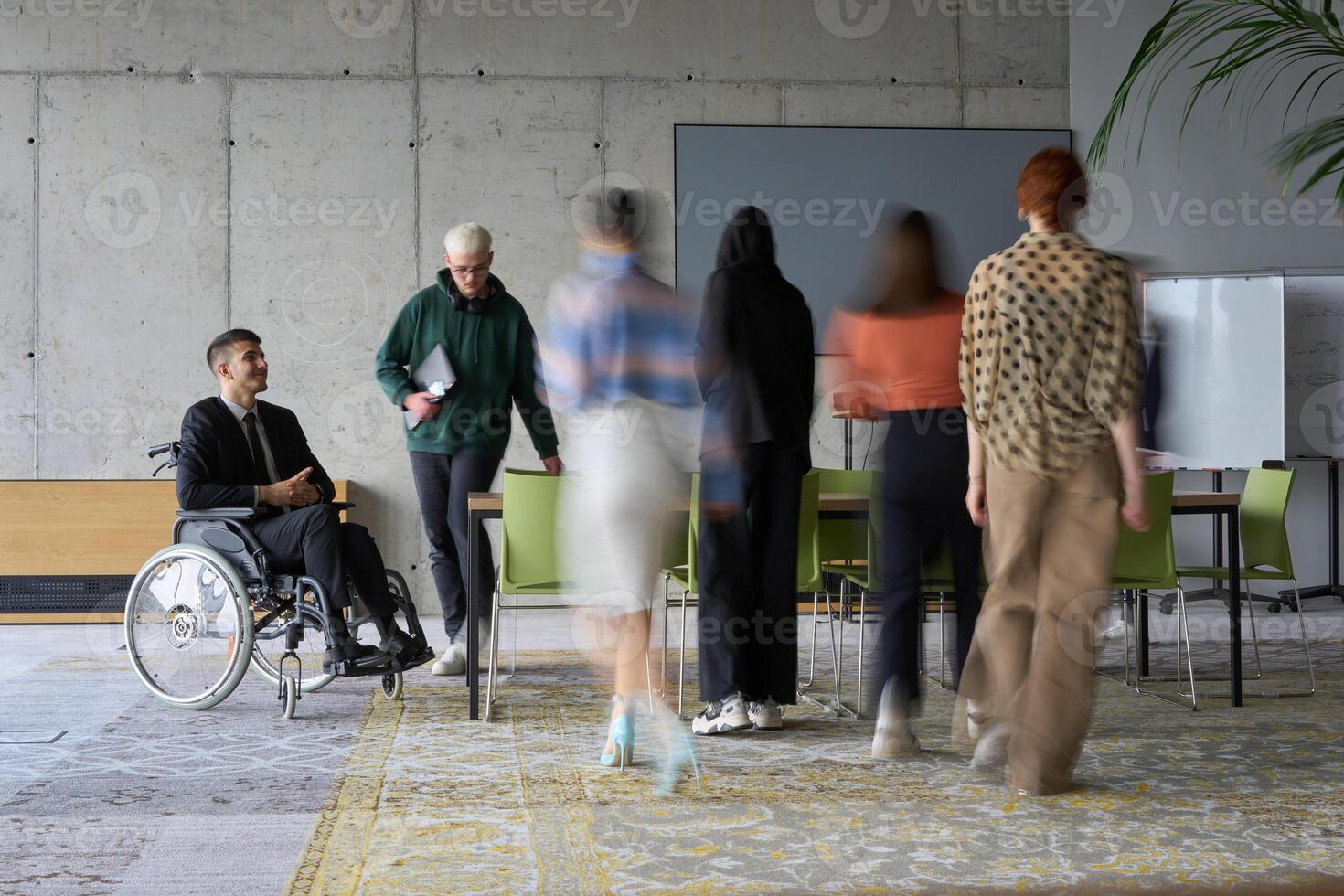 A diverse business team, working urgently around a conference table, collaborates on a meeting while their director, seated in a wheelchair, waits for their input photo